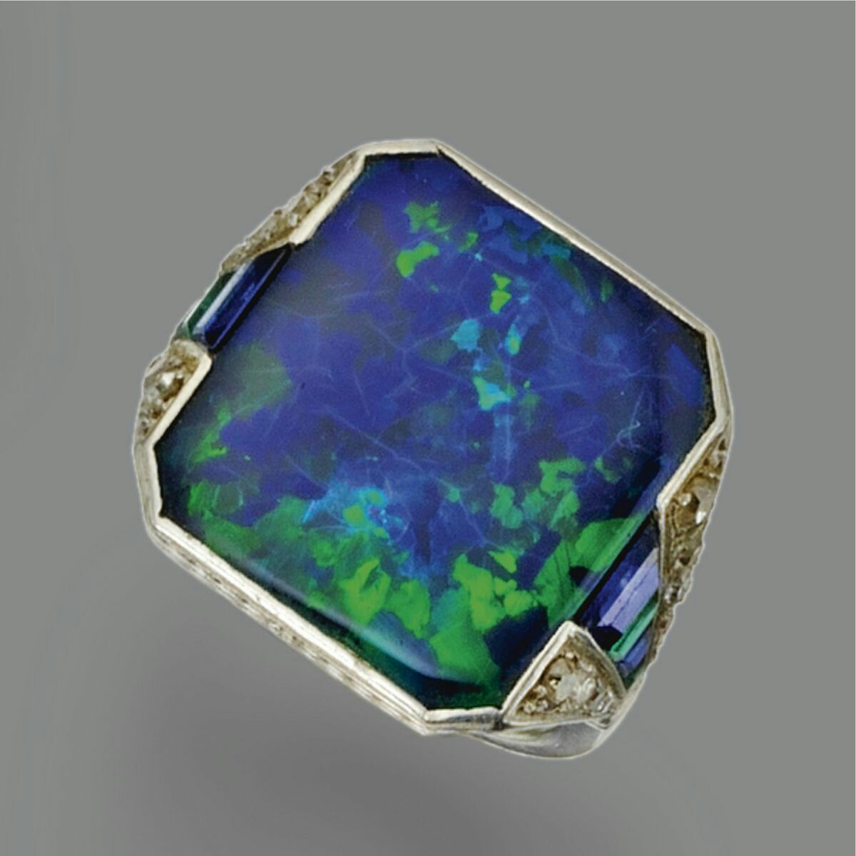 Black opal, colored stone and diamond ring, circa 1930 The black opal of rectangular shape with cut-corner edges measuring approximately 16.5 by 15.0 mm., the shoulders set with tapering bands of calibré-cut emeralds alternating with calibré-cut sapphires within borders of small single-cut diamonds, mounted in platinum, size 5.